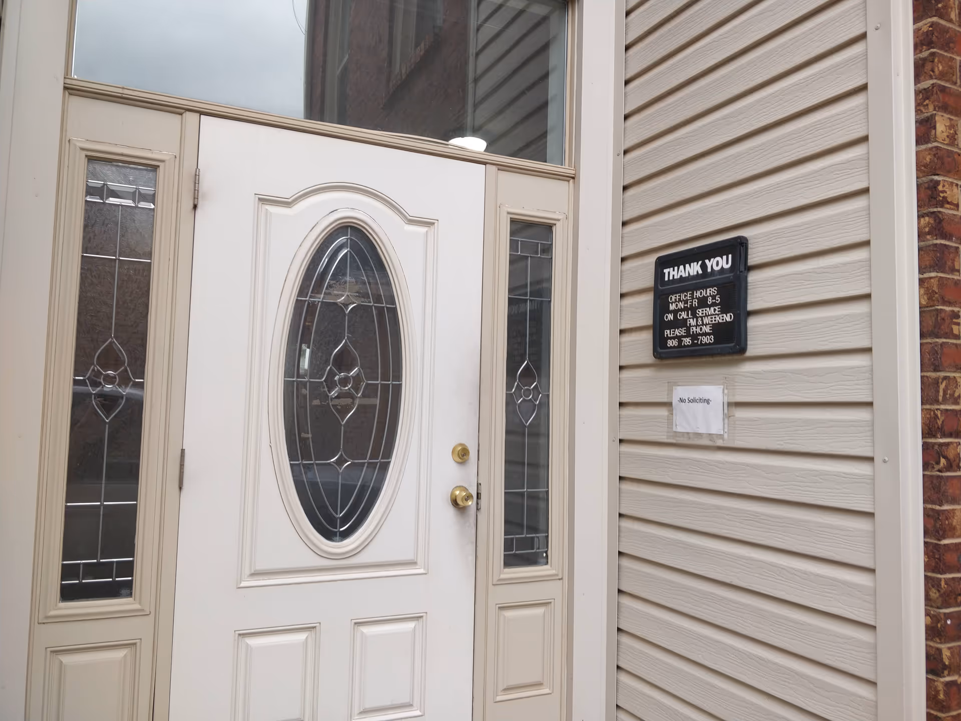 White front door with decorative glass panels, beige siding, and a black hours sign on wall.