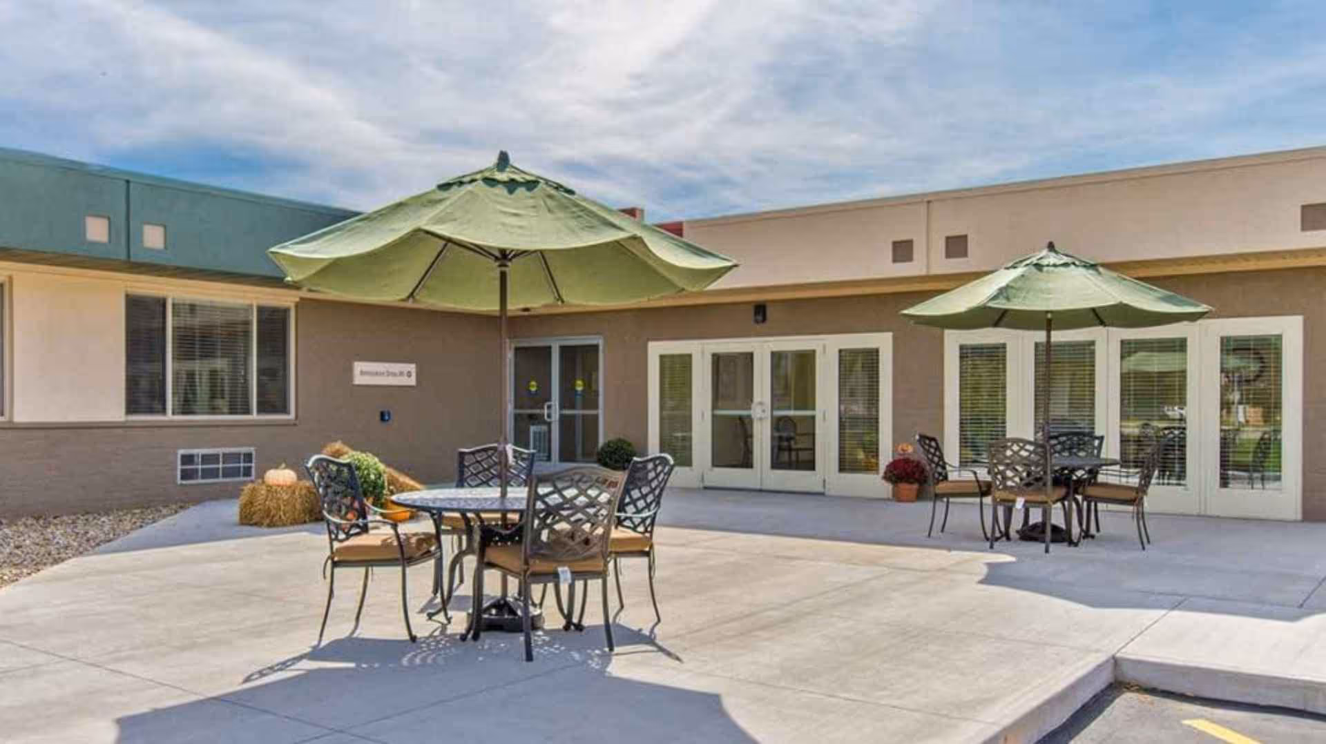 Outdoor patio area at Briarcliff Health & Rehabilitation with two round tables, each surrounded by four chairs and shaded by large green umbrellas. The patio is adjacent to a building with multiple windows and glass doors, under a partly cloudy sky.