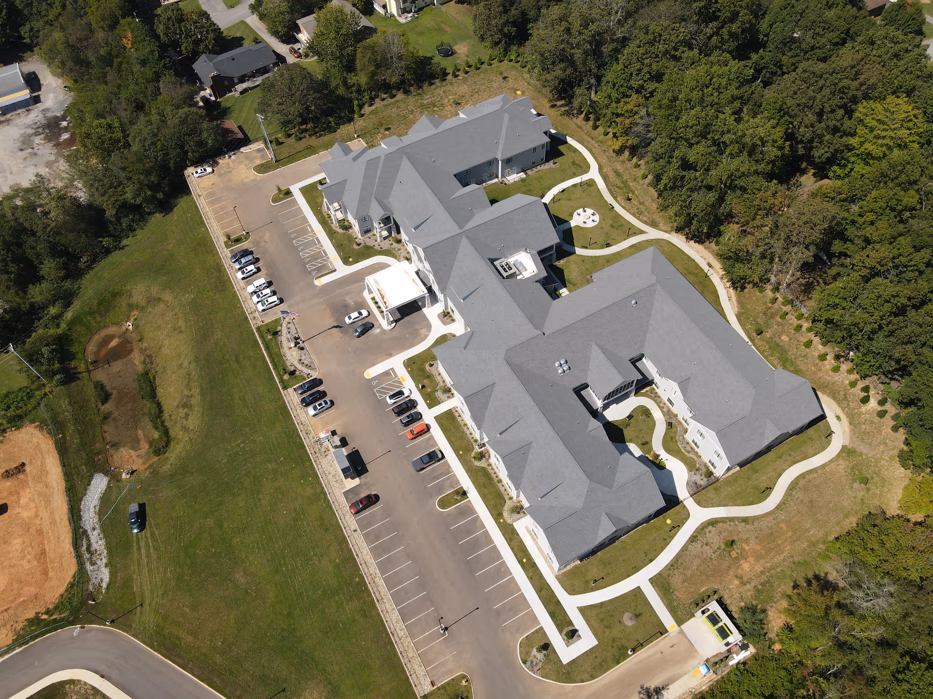 Aerial view of a large senior living campus with grey roofs, parking lot, and surrounding trees.