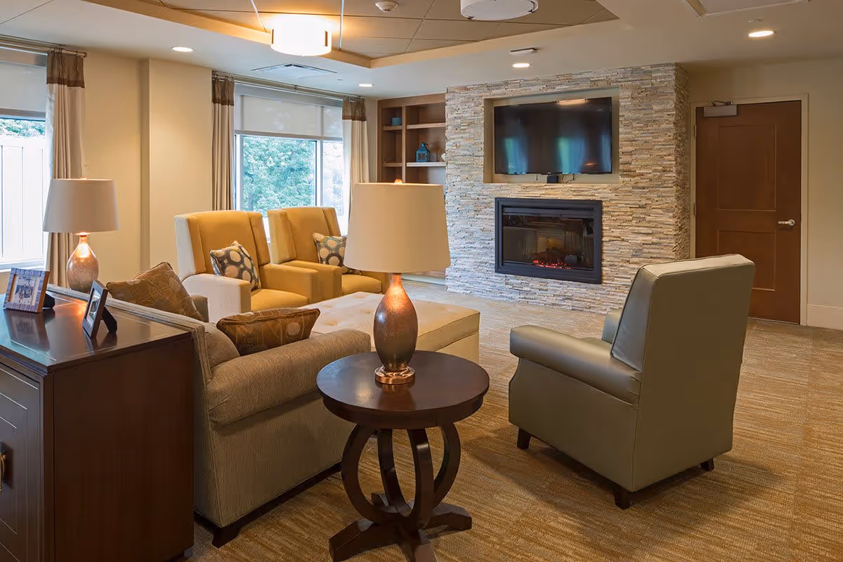 A cozy living room in Silverado Alexandria Memory Care Community featuring comfortable armchairs and a sofa arranged around a round wooden side table with a lamp. The room has large windows with curtains, a stone fireplace with a mounted flat-screen TV above it, and built-in shelves with decorative items.