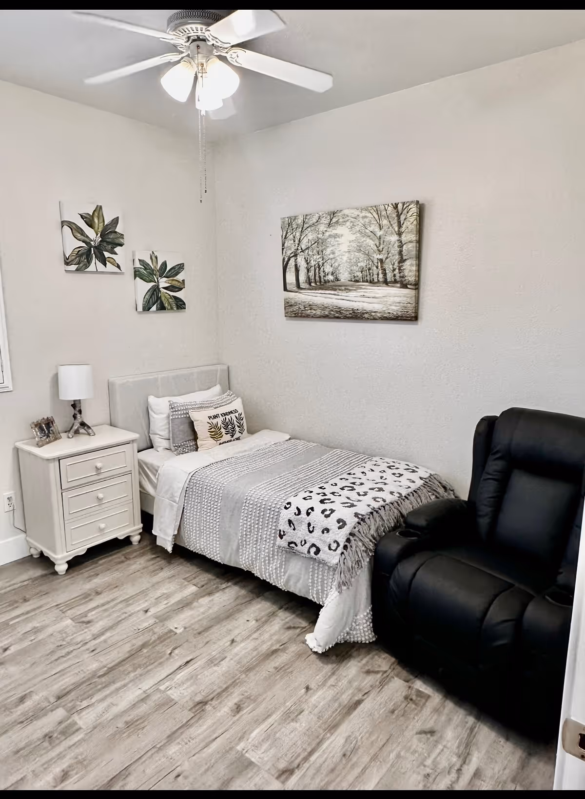 A small, neatly decorated bedroom with a bed, nightstand, lamp, wall art, and a black recliner.