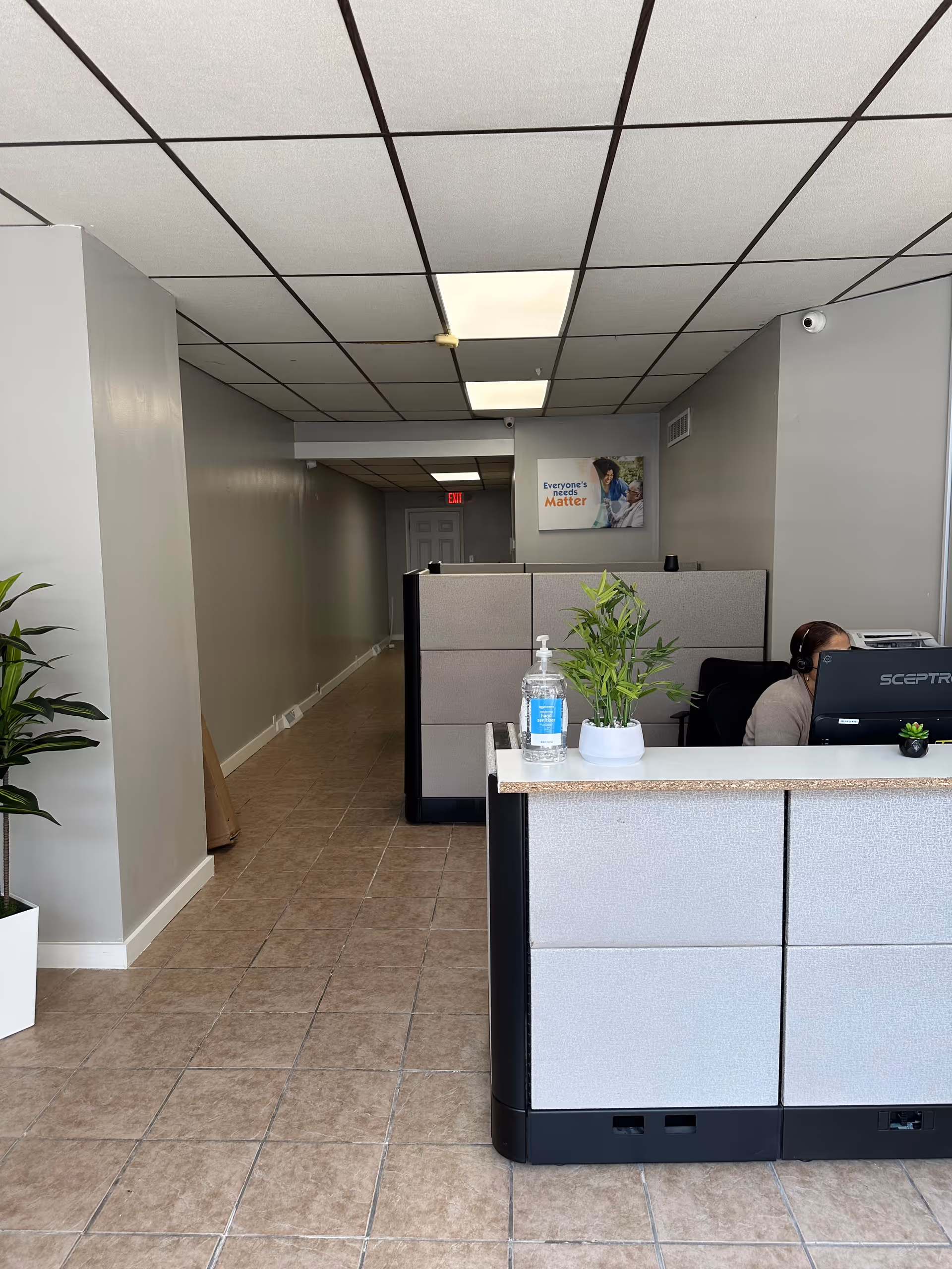 Reception desk with sanitizer, plant, and staff at a computer in a gray-walled lobby.