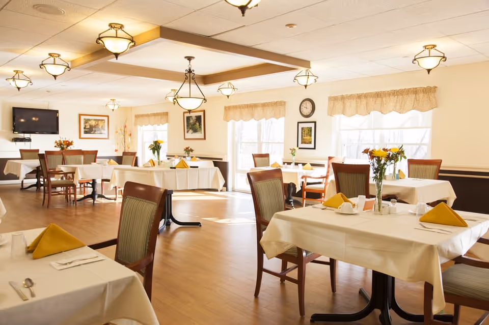 Bright dining room with multiple tables draped in white tablecloths, chairs, yellow folded napkins and floral centerpieces.