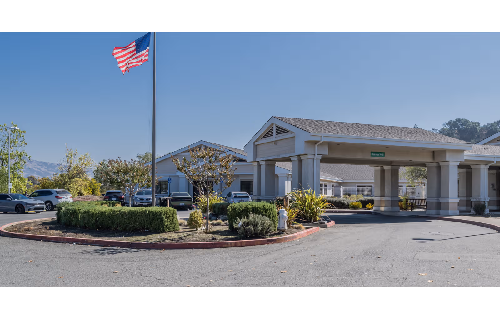 Wide view of Tice Valley Post Acute's entrance with flag, parked cars, and blue sky.
