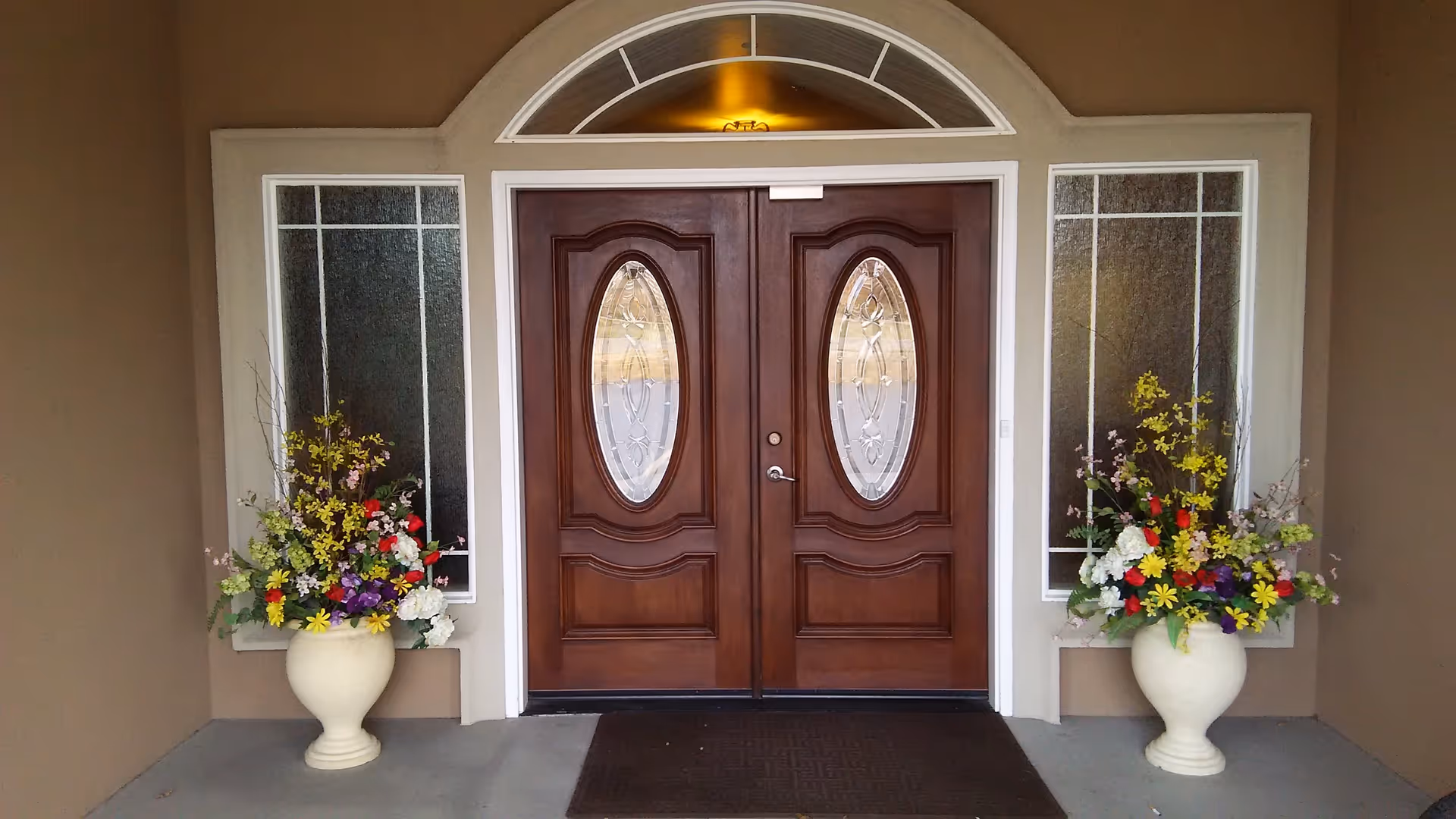 Front entrance of a building with double wooden doors featuring oval glass panels. On each side of the doors are tall, narrow windows with textured glass. Two large cream-colored vases filled with colorful artificial flowers are placed symmetrically on either side of the entrance. A dark doormat lies in front of the doors.