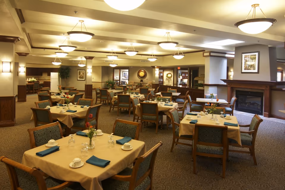 Spacious dining room with multiple tables set with napkins, cups and flowers, warm lighting, and a fireplace.