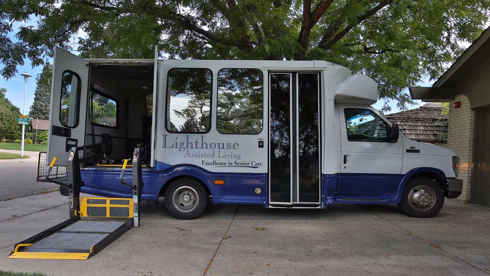 White and blue Lighthouse Assisted Living shuttle bus parked outside the facility with a ramp.