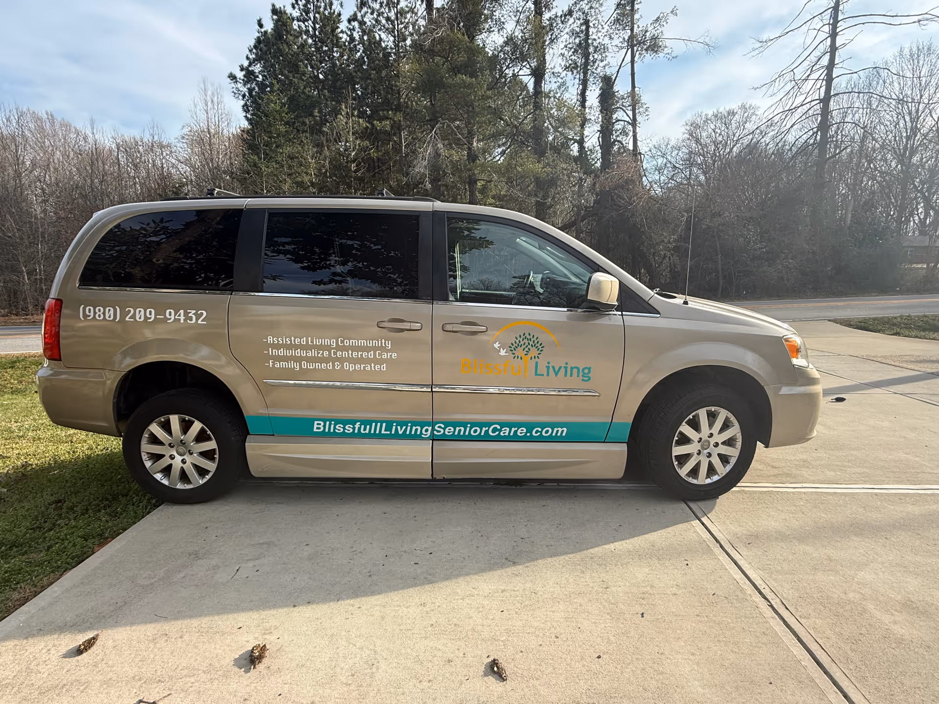 Beige Blissful Living van parked outdoors displaying logo, contact number, and website on its side.