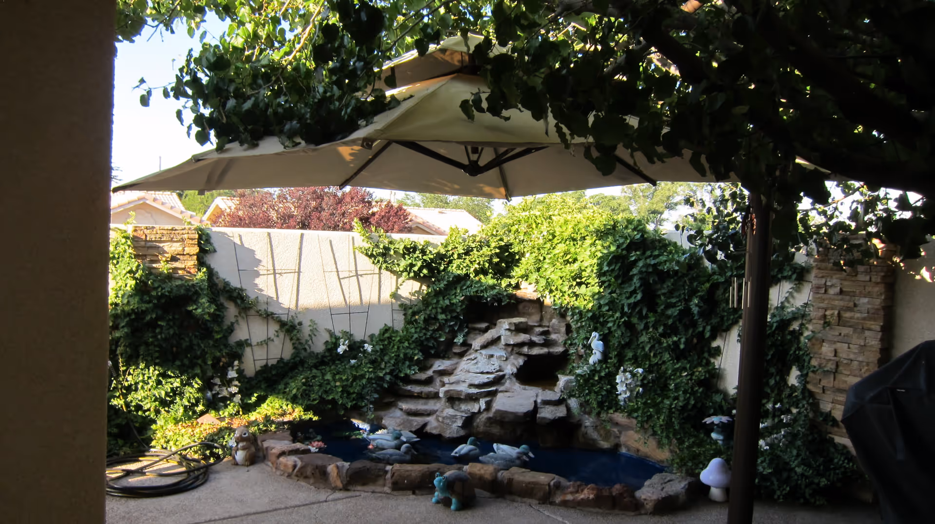 A shaded outdoor patio area with a large umbrella, surrounded by lush green ivy and plants. There is a small rock waterfall feature with a pond at the base, decorated with small animal figurines. The area is enclosed by a beige wall and has a stone pillar on the right side.