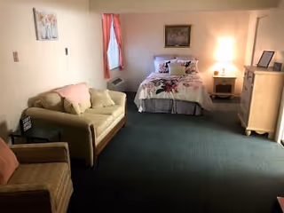 Studio-style room with a bed, sofa, armchair, dresser, nightstand with a lamp, and a window with pink curtains.