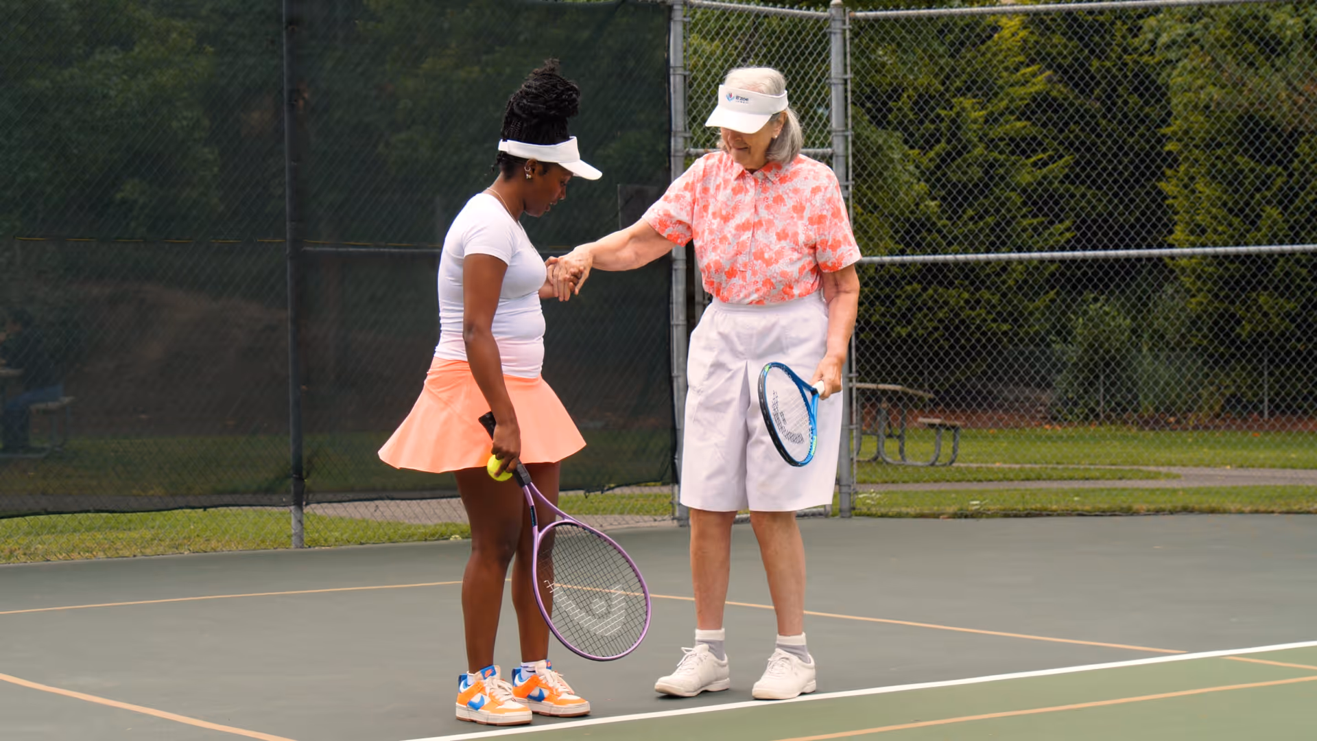 Two women on a tennis court shake hands after a game, holding rackets.