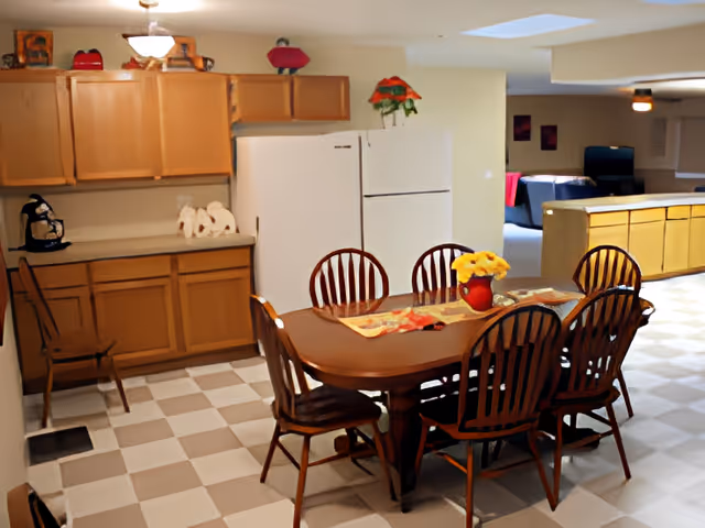 Dining area with a wooden table and six chairs next to kitchen cabinets and two refrigerators in a communal room.