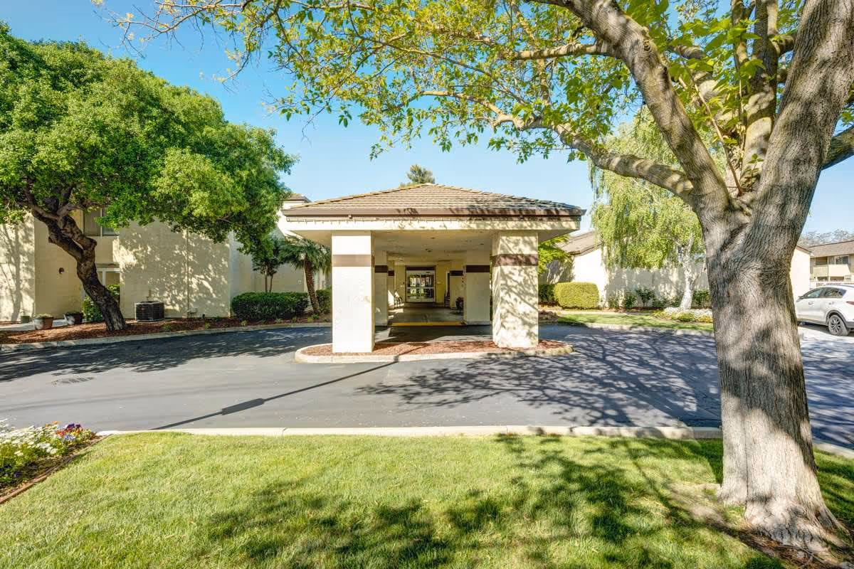 Exterior view of The Oaks at Inglewood facility entrance with a covered driveway, surrounded by green trees, grass, and a clear blue sky.