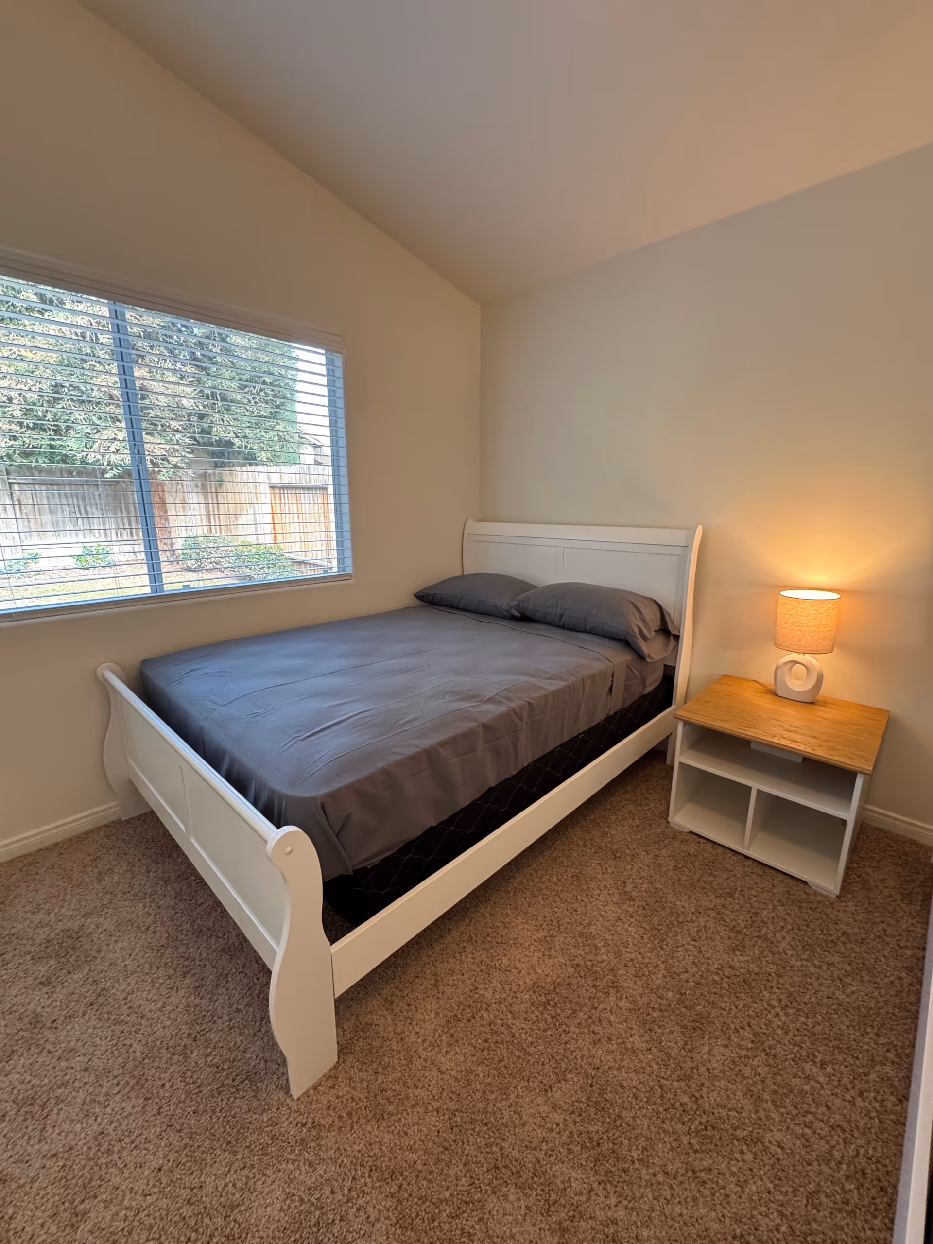 White bed with gray bedding and a nightstand sits in a small beige senior living bedroom