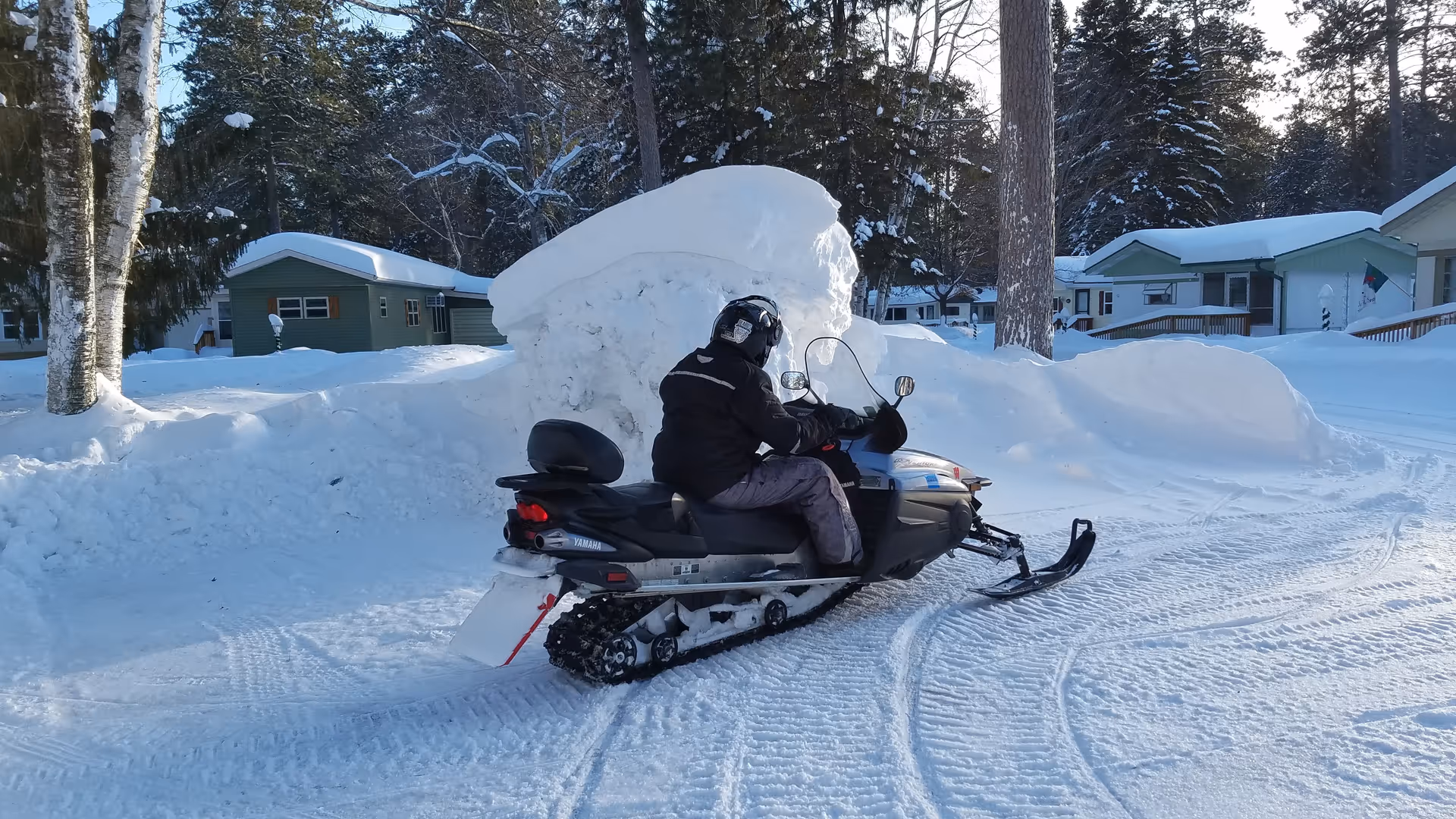 Person riding a snowmobile along a snow-covered residential street with large snowbanks.
