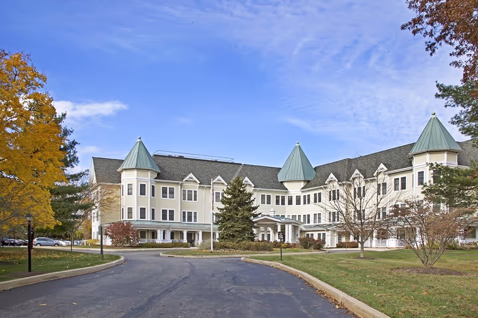 Exterior view of a large, three-story senior living facility with light-colored walls and green peaked roofs. The building is surrounded by trees with autumn foliage and a paved driveway leading to the entrance.