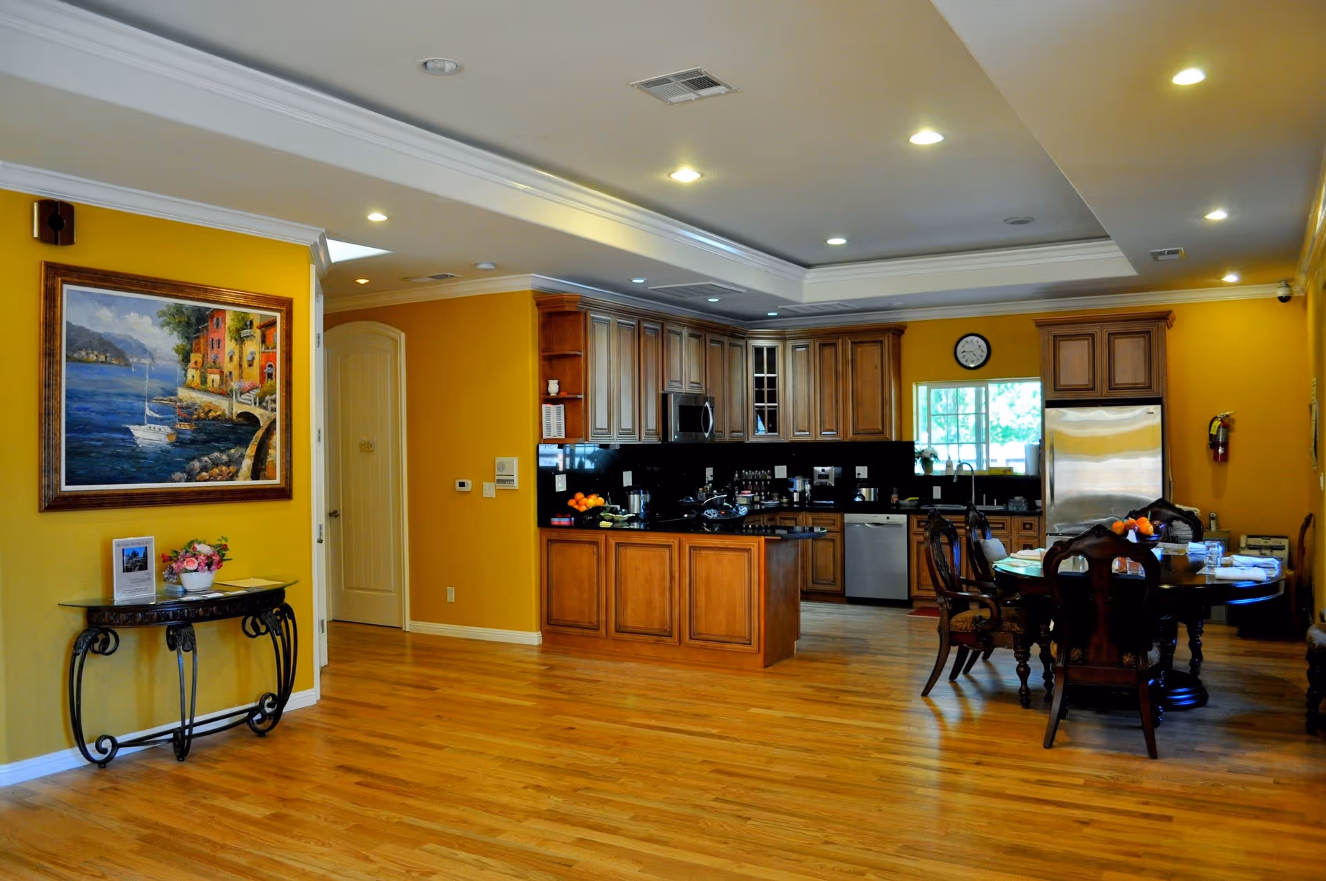 Open-concept kitchen with wooden cabinets, island, dining table, and yellow walls in a senior living home.