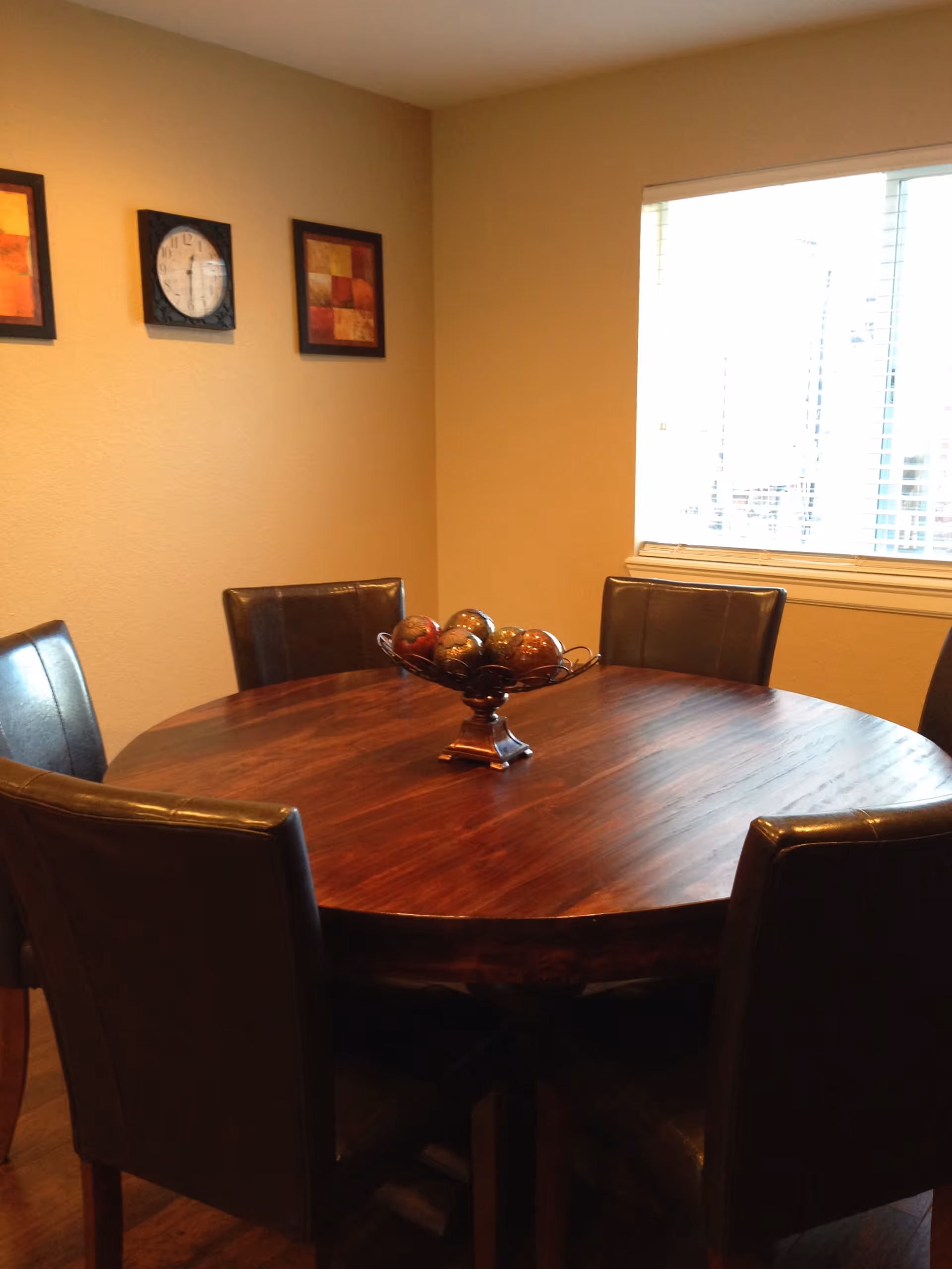 A round wooden dining table surrounded by six dark leather chairs in a room with beige walls. On the table is a decorative bowl filled with ornamental balls. A window with blinds is on the right wall, and two framed pictures and a clock hang on the left wall.