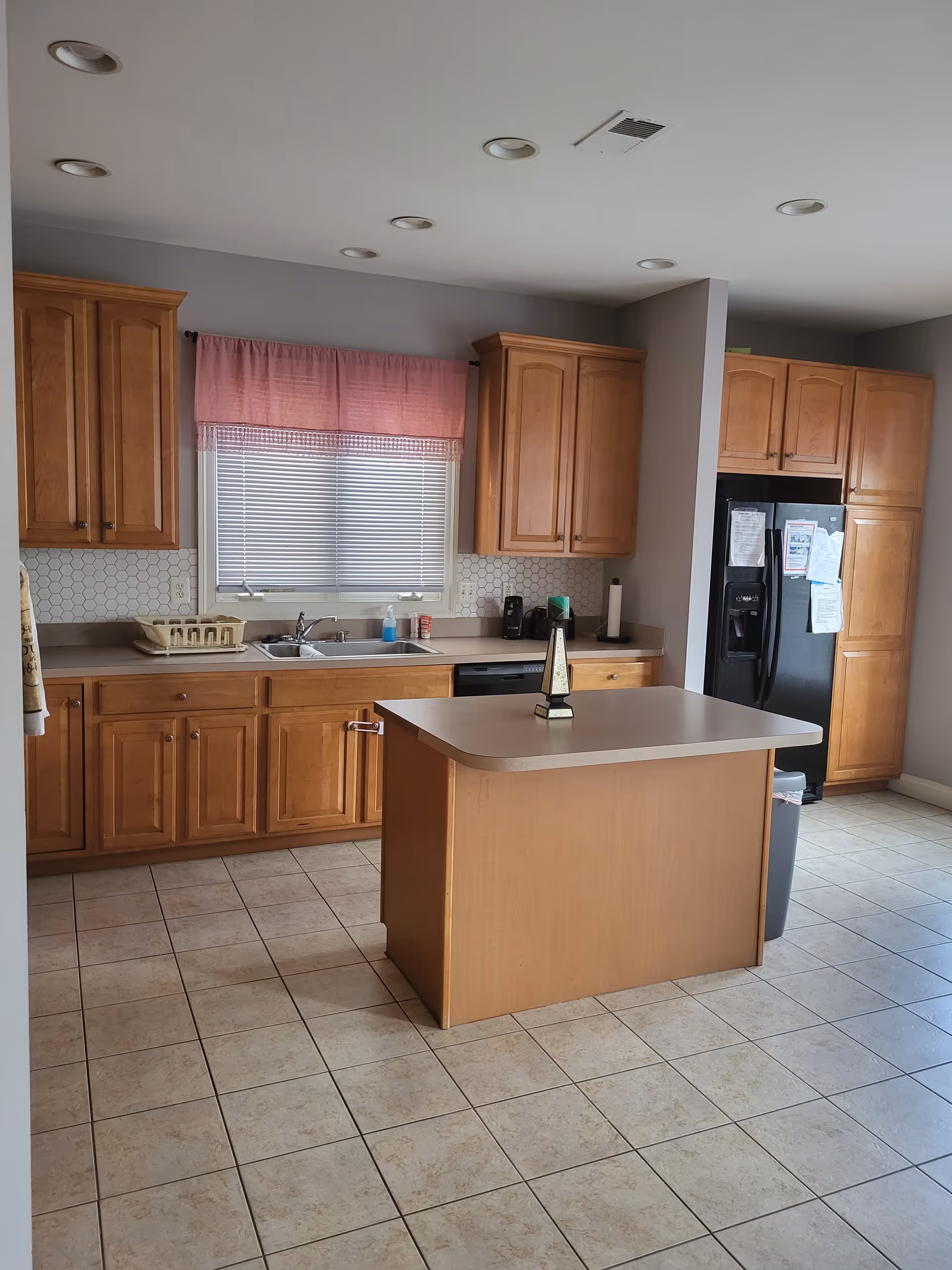 Wooden kitchen cabinets surround a central island in a bright kitchen with sink and refrigerator.