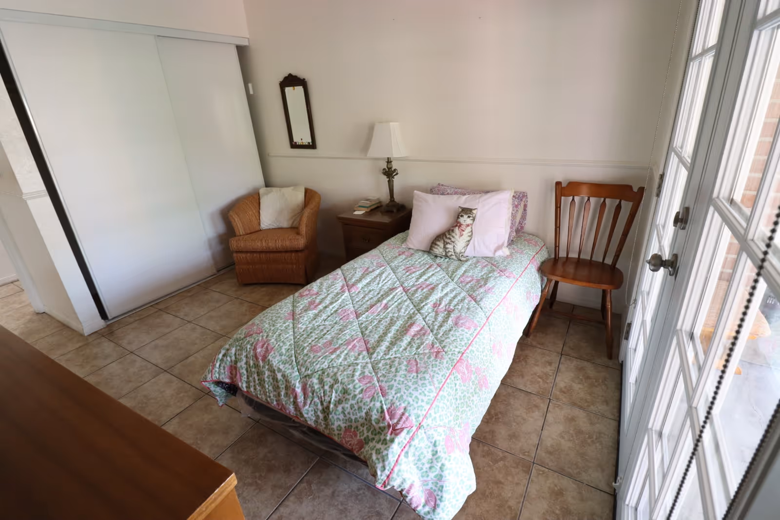 Small bedroom with floral quilted bed, nightstand, two chairs, and large glass doors.