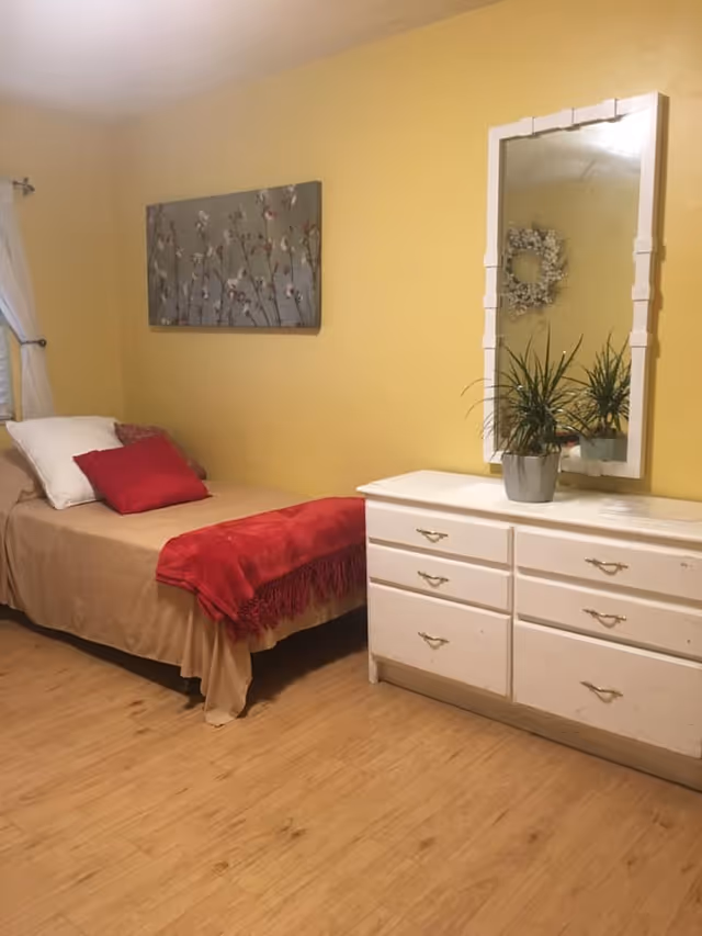 Cozy bedroom in a senior living facility with a bed, dresser, mirror, and plants.