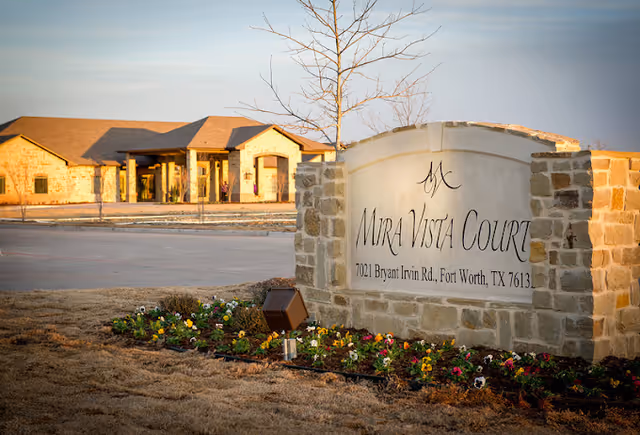 Stone entrance sign reading "Mira Vista Court" with flowers in front of a single-story building under a clear sky.