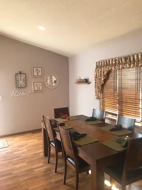 A dining room with a wooden table set for six people with green placemats and folded napkins. The room has wooden flooring, a window with wooden blinds and floral valance, and a light purple wall decorated with a clock, framed pictures, a round mirror, and the word 'blessed'.