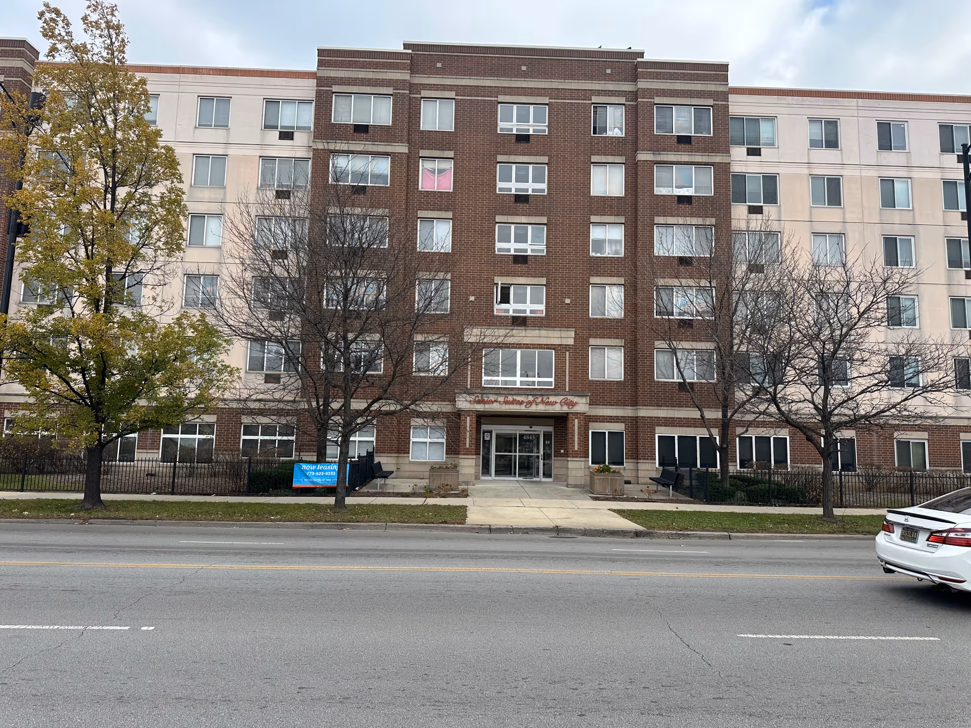 Front exterior of a multi-story brick and stucco senior living building with central entrance, sidewalk and trees.