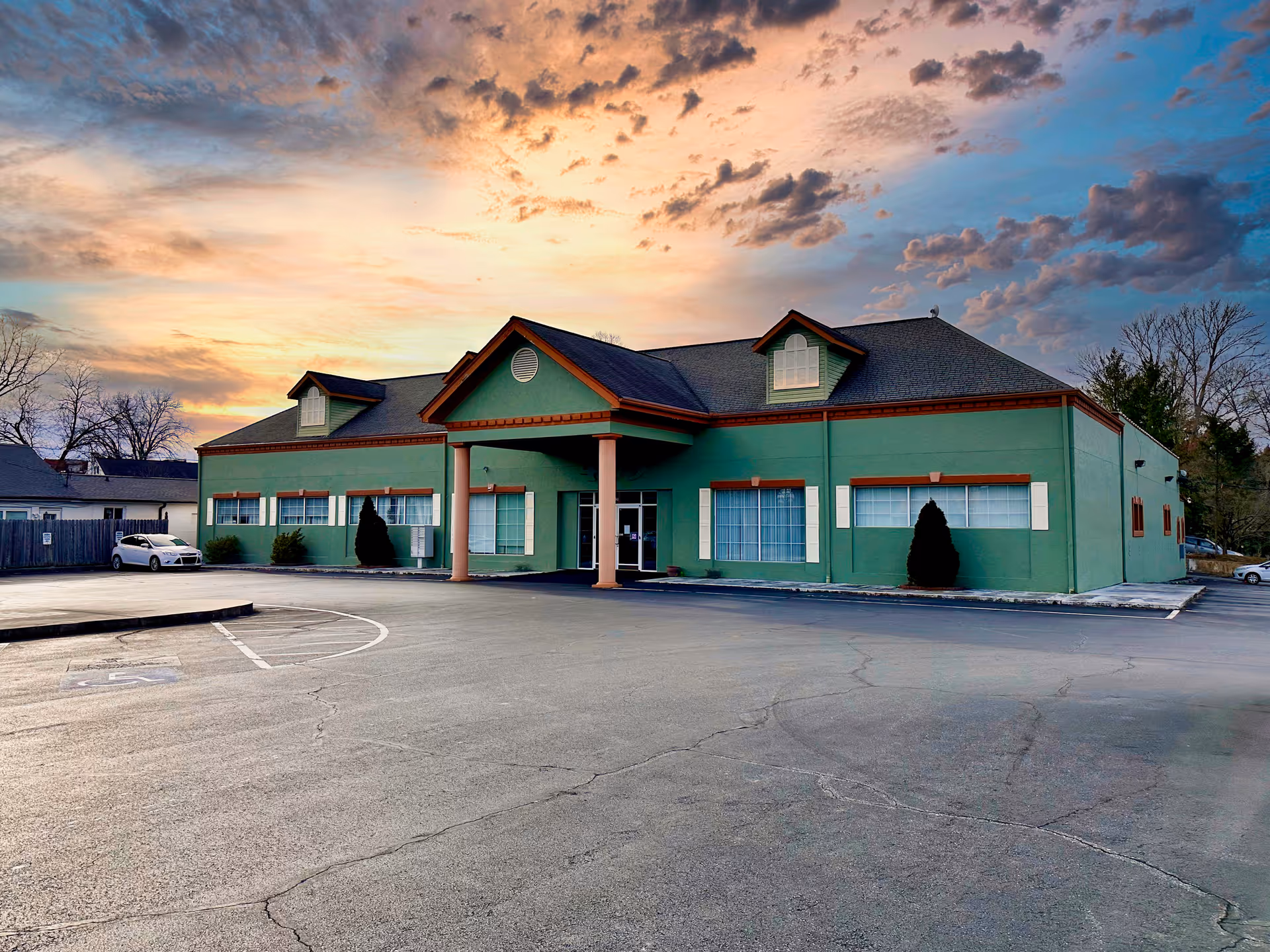 Green healthcare facility front with covered entrance and surrounding parking lot.