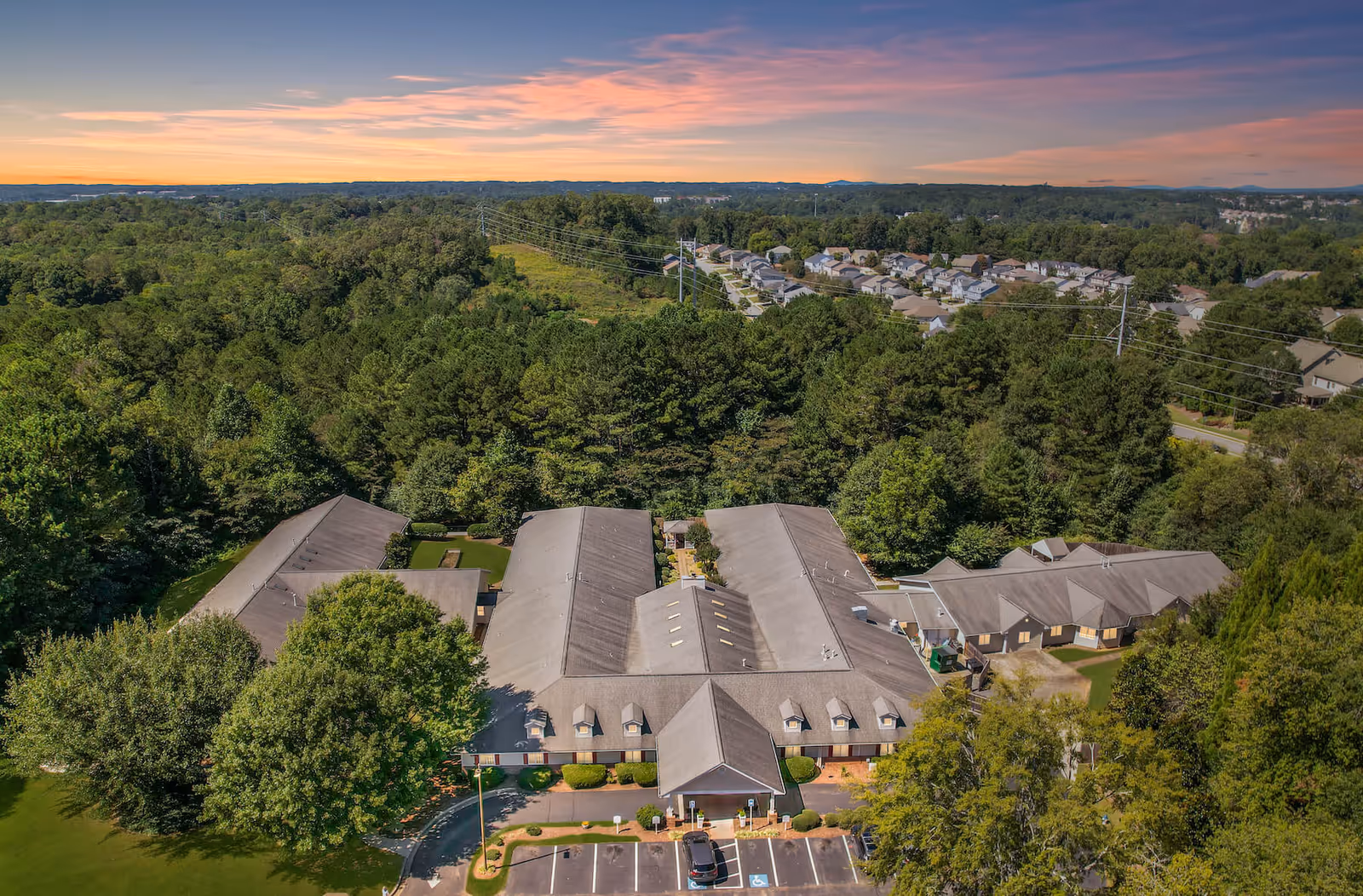 Aerial view of Bellevue Village at Neese Road senior living facility surrounded by dense green trees and residential houses in the distance under a colorful sunset sky.