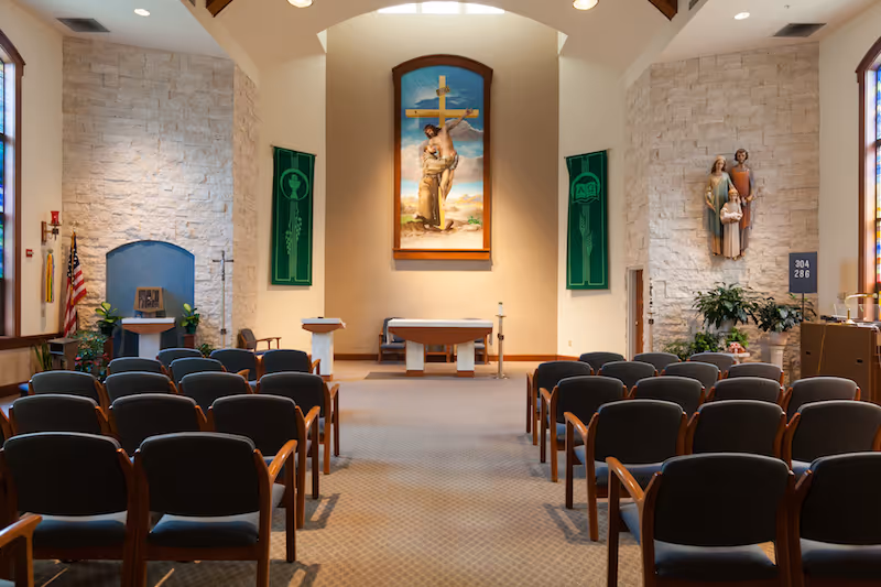 Interior view of a chapel featuring a large cross painting, seating arrangement, and decorative elements including plants and religious statues.
