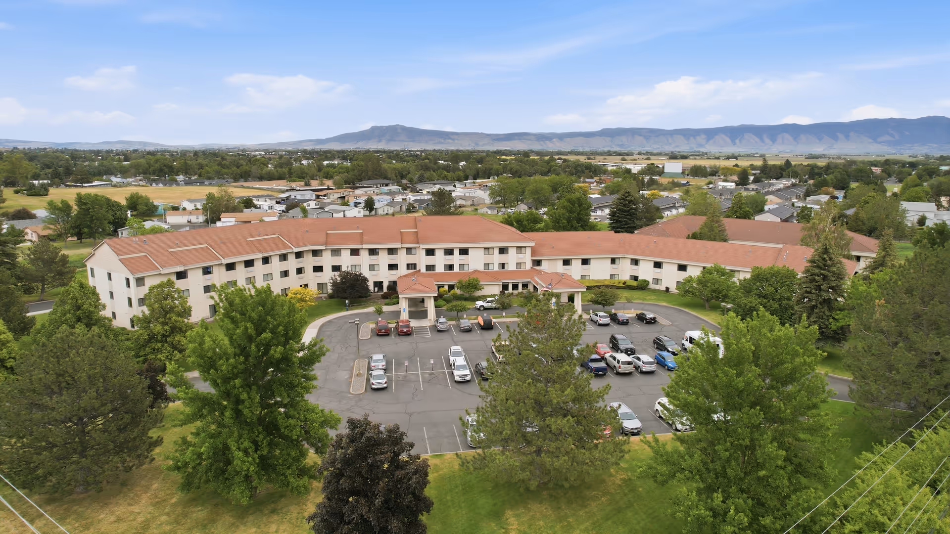 Aerial view of a large retirement residence with red roof, parking lot, and surrounding trees.