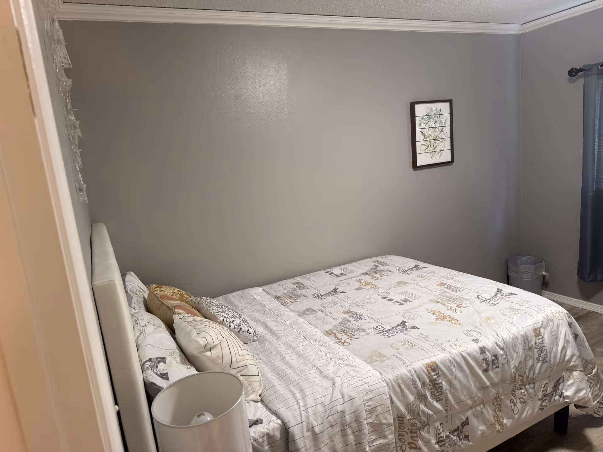 Small furnished bedroom with a bed, pillows, nightstand, lamp, and framed wall art.