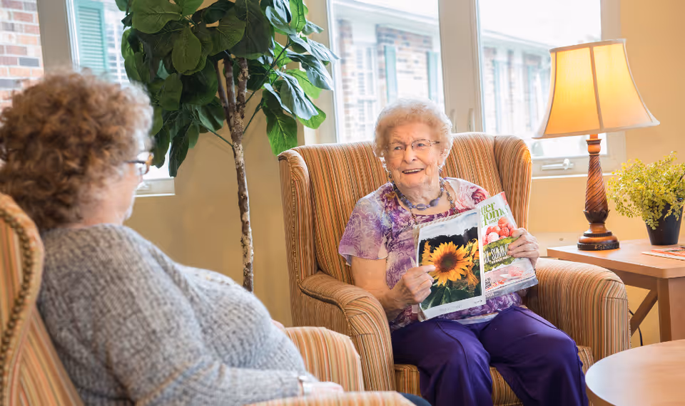 Two elderly women sitting in comfortable armchairs in a well-lit room. One woman is holding and showing a magazine with a sunflower image on the cover, while the other woman listens attentively. There is a tall green plant near the window, a table with a lamp and a small potted plant beside them.