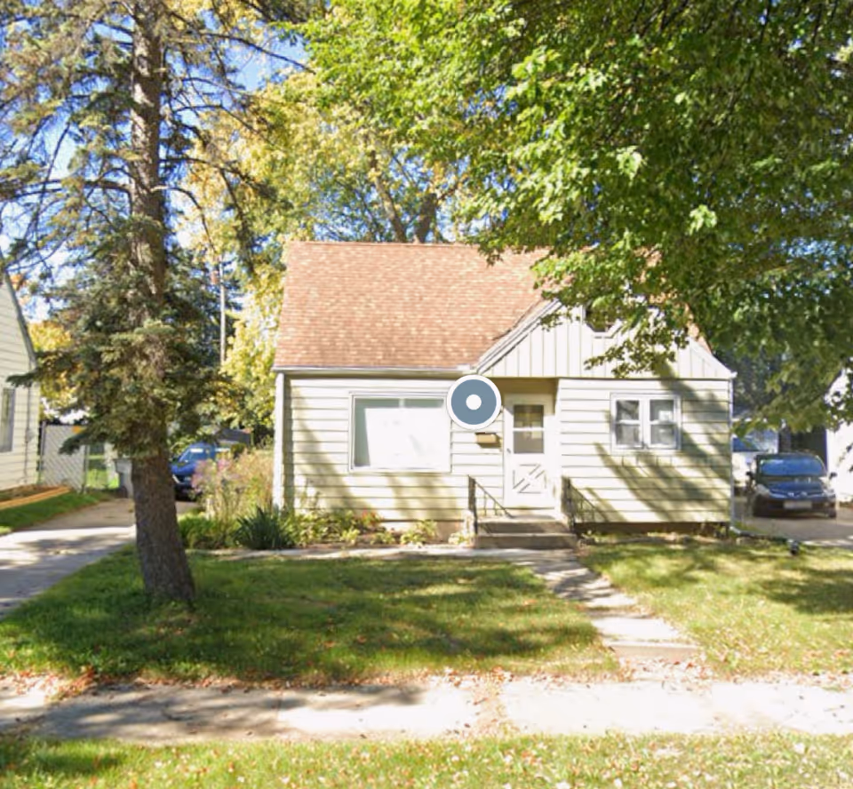 Front exterior of a small beige house with a front door and surrounding trees.