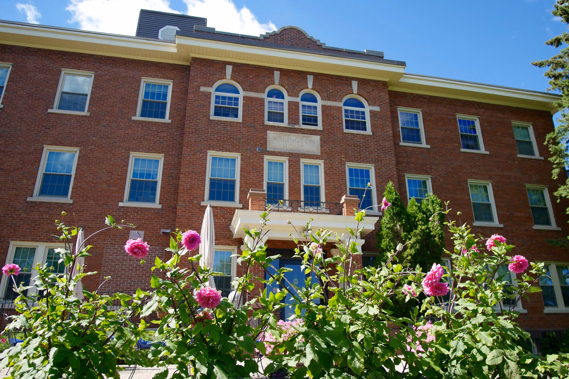 Exterior view of a three-story brick building with multiple windows and a small balcony above the entrance. In the foreground, there are green plants with pink flowers. The sky is clear and blue with some clouds.