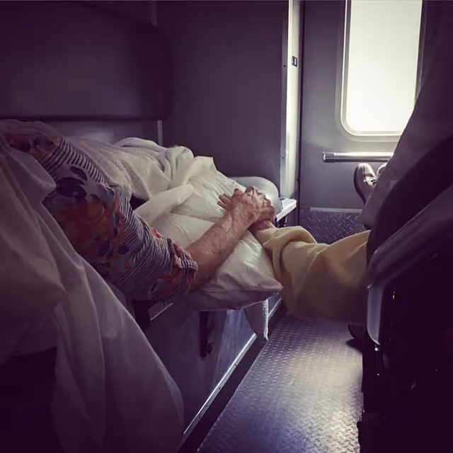 An elderly patient lies in bed while a caregiver holds their hand.