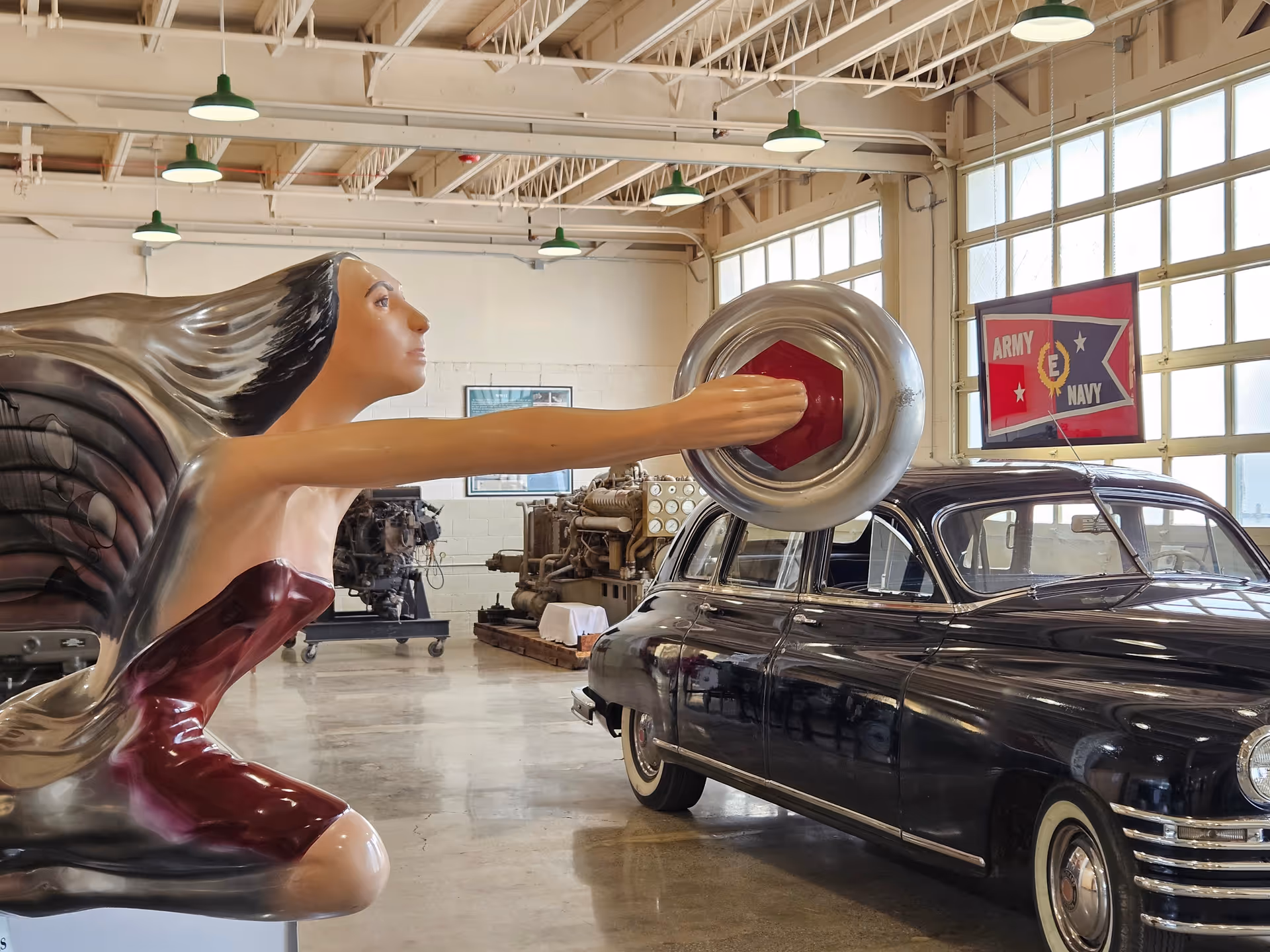 A large indoor showroom features a vintage car and a giant sculpture reaching toward the vehicle.