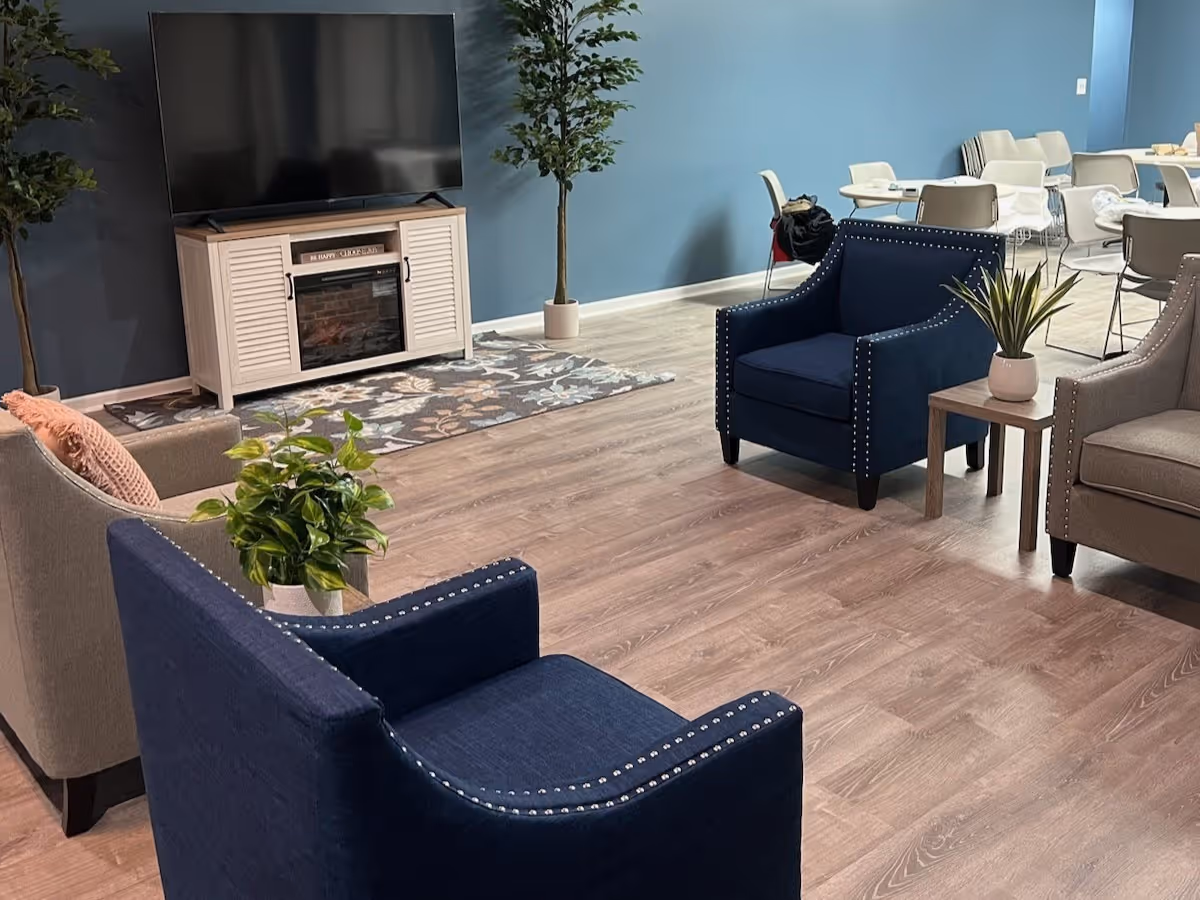Modern senior living lounge with blue upholstered chairs, plants, and a TV on a stand.