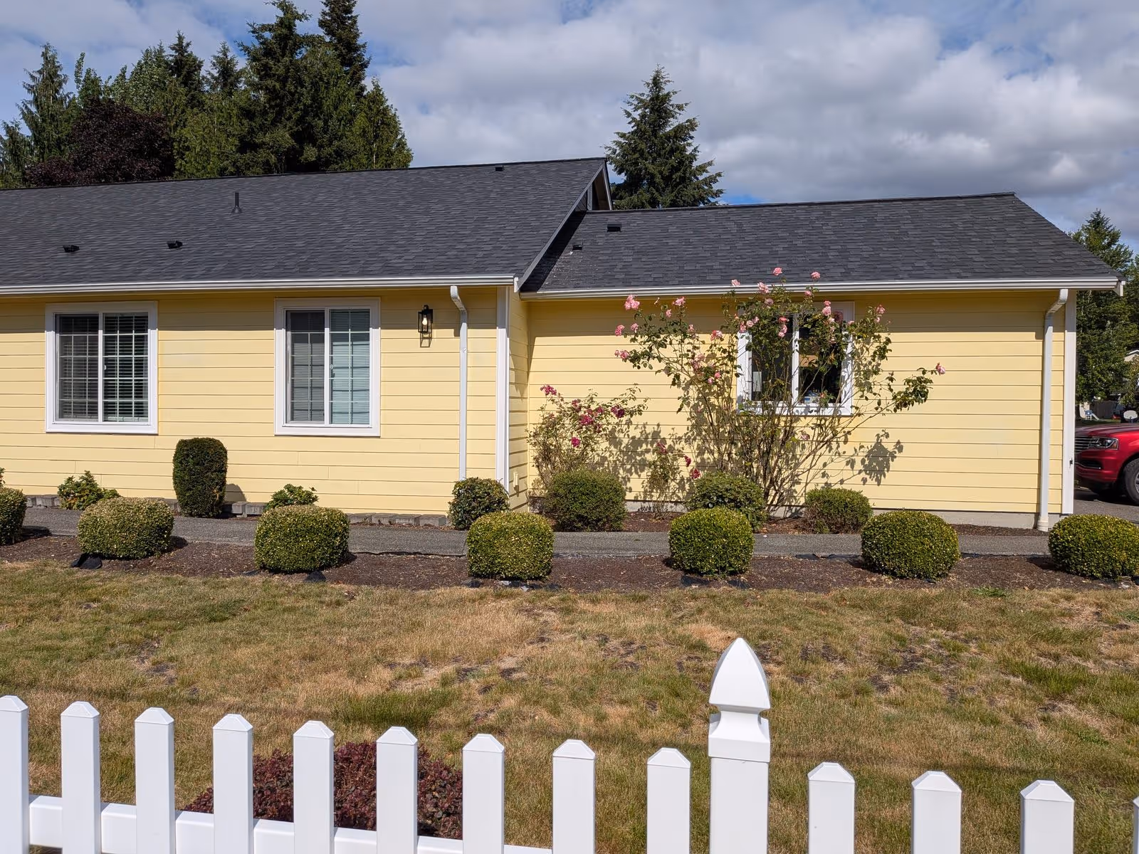Single story yellow house exterior with white trim, trimmed shrubs, and a white picket fence.