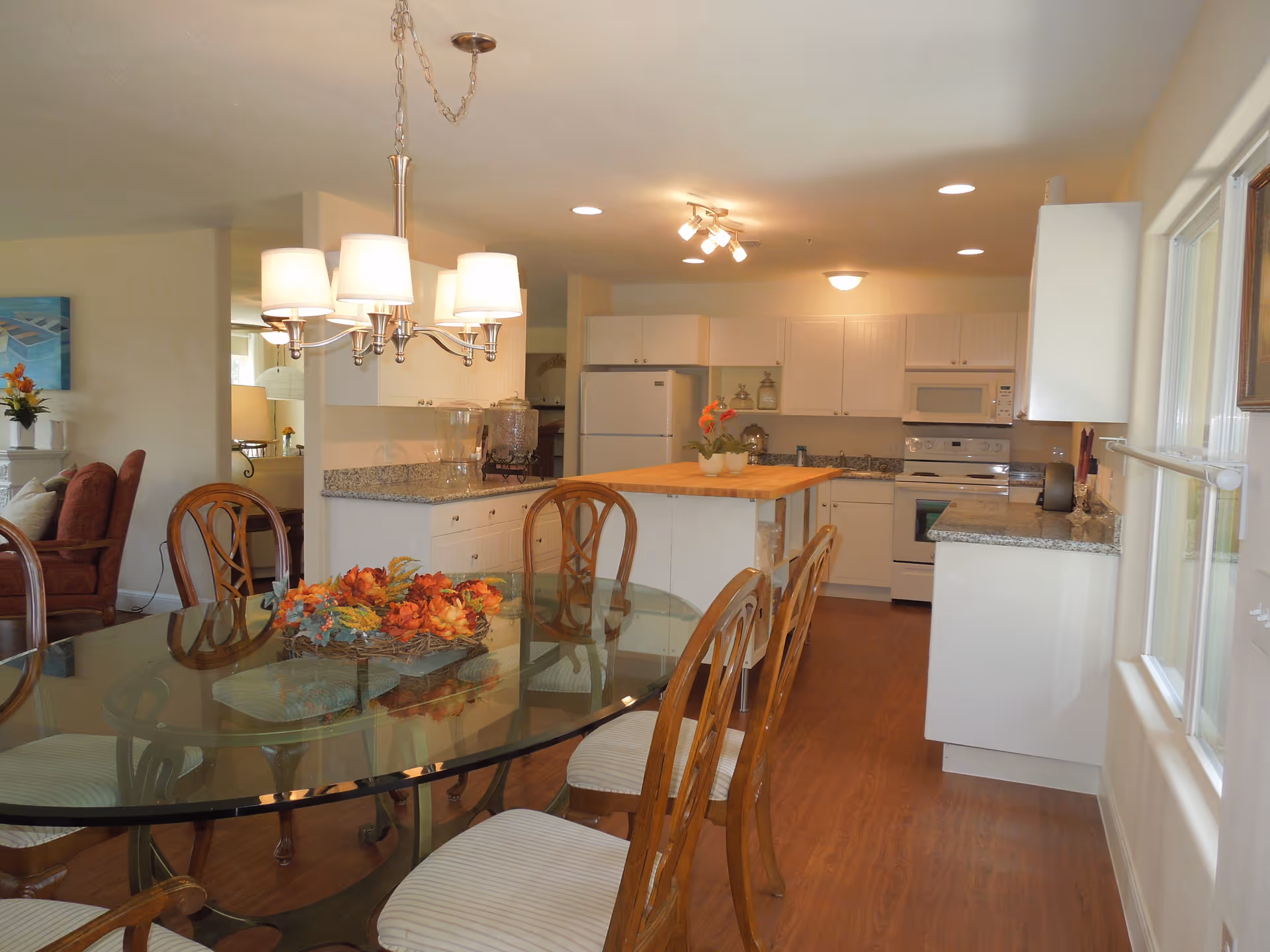 Interior view of a dining area and kitchen in a senior living facility. The dining area features a glass-top table with six wooden chairs, decorated with a floral centerpiece. The kitchen has white cabinets, granite countertops, a white refrigerator, stove, microwave, and a wooden island with small potted plants. The room has wooden flooring and is well-lit with ceiling lights and a chandelier above the dining table.