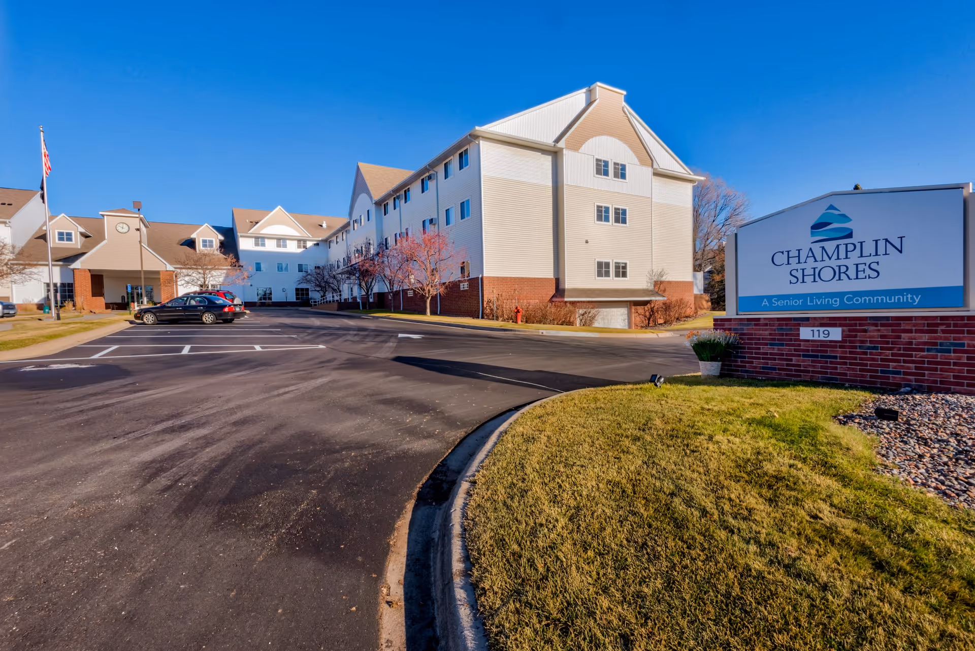 Front exterior of the Champlin Shores senior living community building with a driveway, parking area, and entrance sign.
