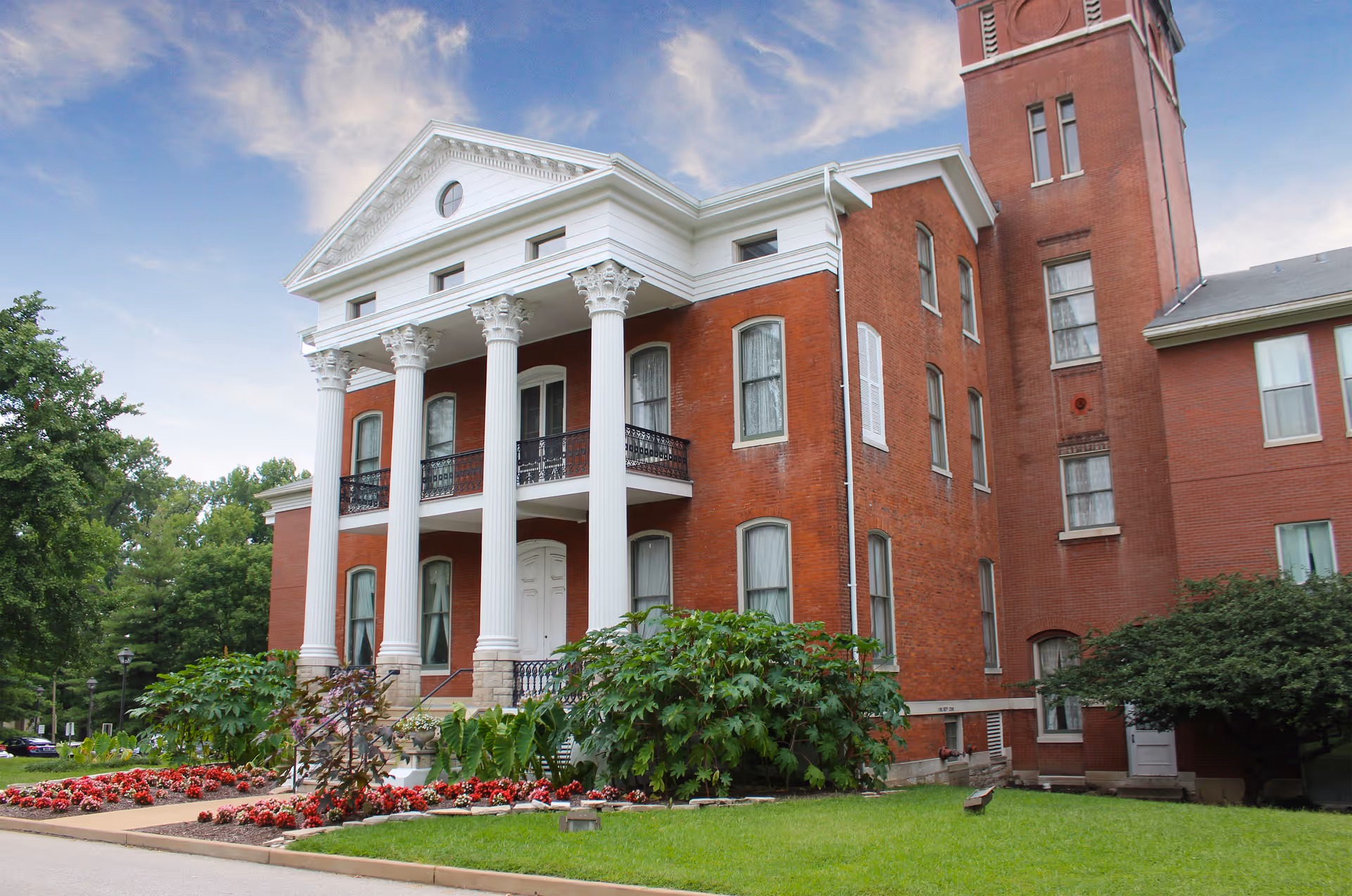 Front view of a red-brick historic building with white columns, balconies, and landscaped grounds.