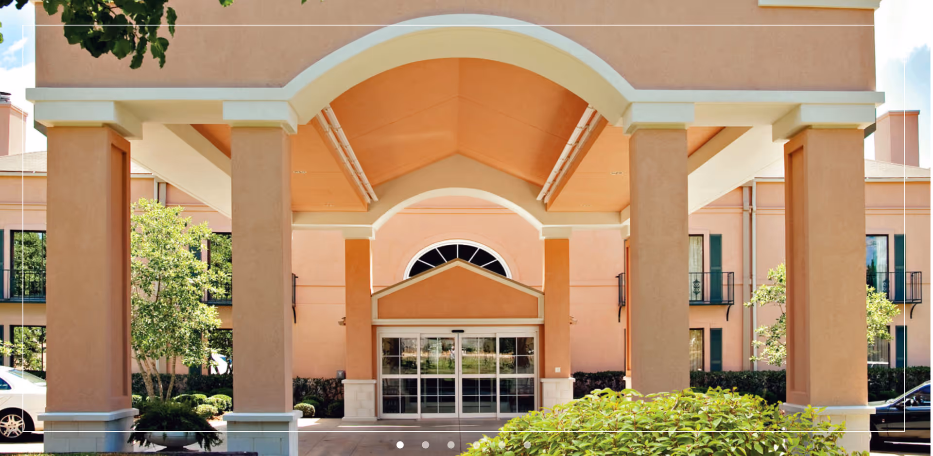 Entrance of a senior living facility named The Orchard, featuring a covered driveway with large peach-colored pillars and a glass door entrance. The building has multiple windows and is surrounded by greenery and trees.