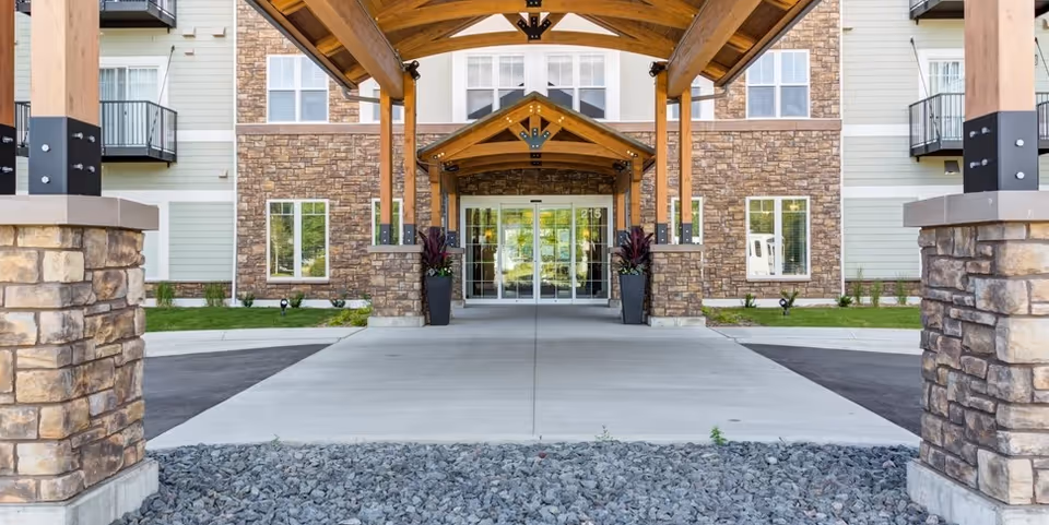 Front entrance of a senior living building featuring stone columns, wooden canopy, and glass doors.