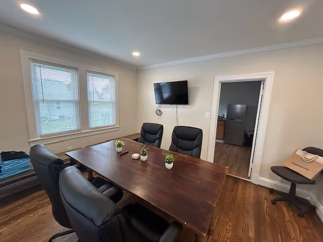 Conference room with a long wooden table, black chairs, plants, windows, and a wall-mounted TV.