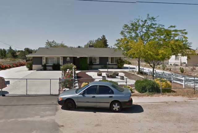 One-story care facility exterior with a gated driveway, shrubs, and a parked car on the street.