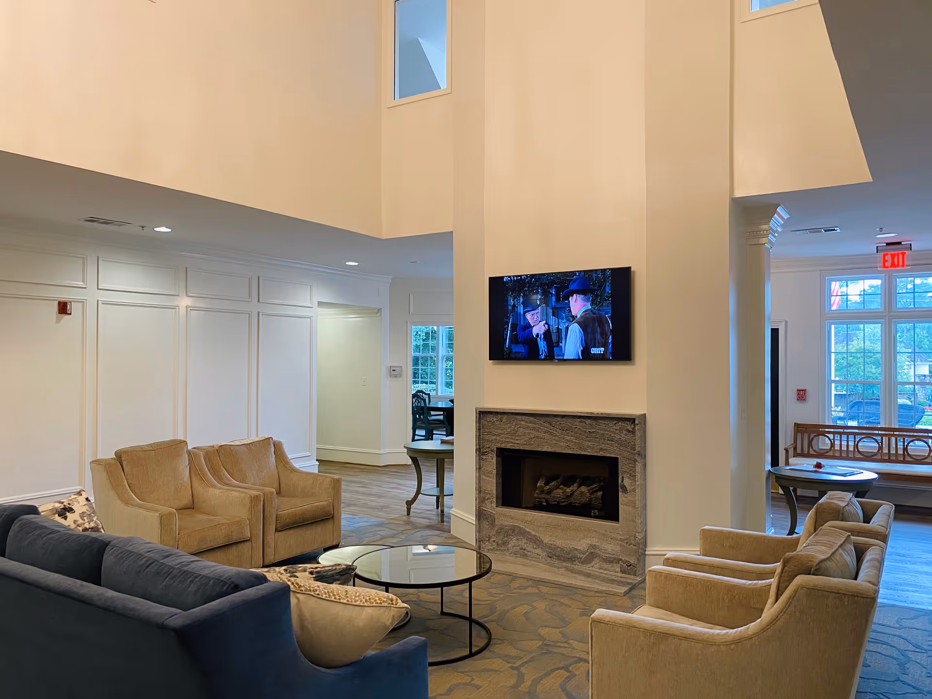 A cozy senior living facility common area with a fireplace and a wall-mounted TV above it. The room features comfortable seating including a blue sofa, beige armchairs, and a glass coffee table. Large windows allow natural light to fill the space, and there is a wooden bench near the entrance with an exit sign above the door.