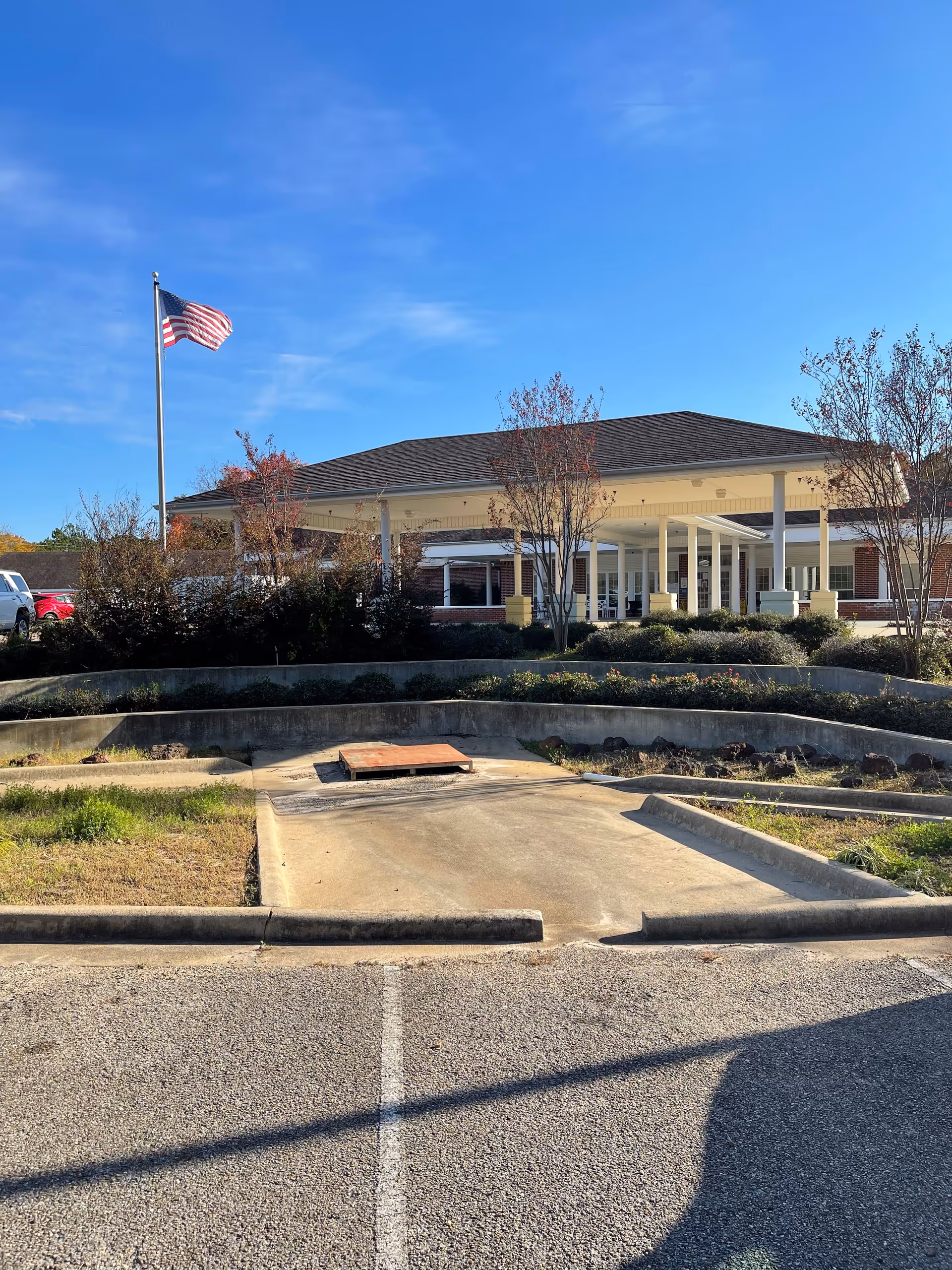 Front entrance of a single-story nursing home building with a covered porte-cochère, landscaping, and an American flag on a flagpole.