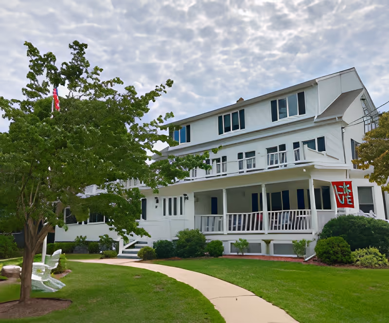 Three-story white building with a wide covered front porch, pathway, green lawn and trees under a cloudy sky.